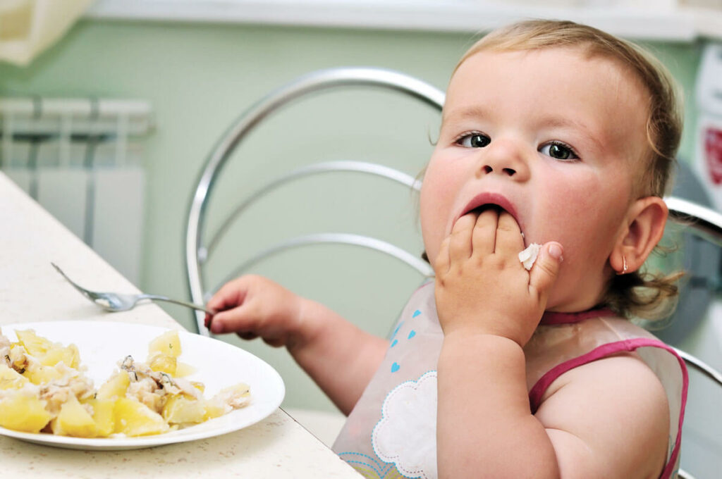 Toddler eating potatoes with fingers at table.