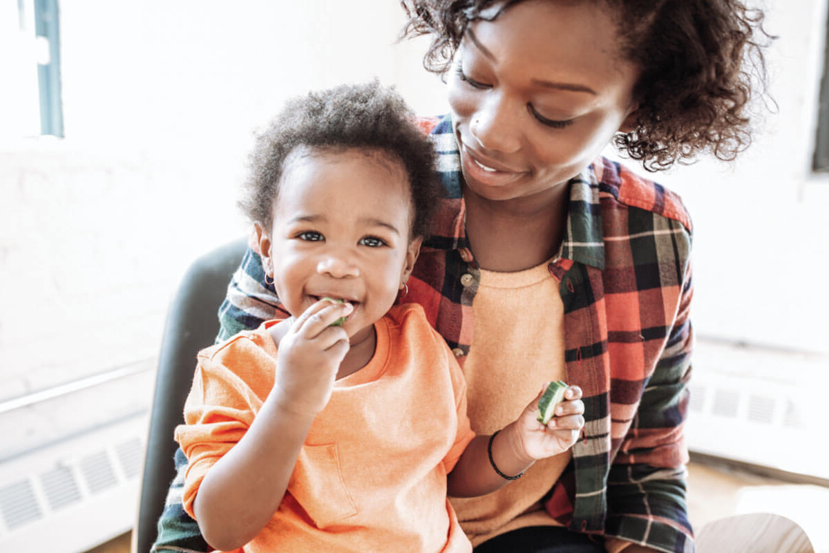 Child eating cucumber on woman's lap, both smiling.