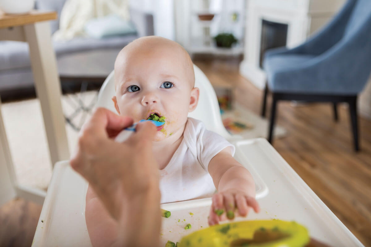 Baby being spoon-fed green puree in high chair.
