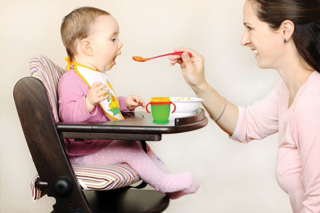 Caregiver feeds baby in high chair with spoon.