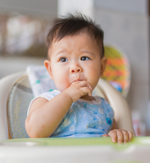 Baby chewing fingers in high chair, looking curious.