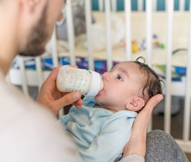 Parent feeding baby with a bottle near crib.