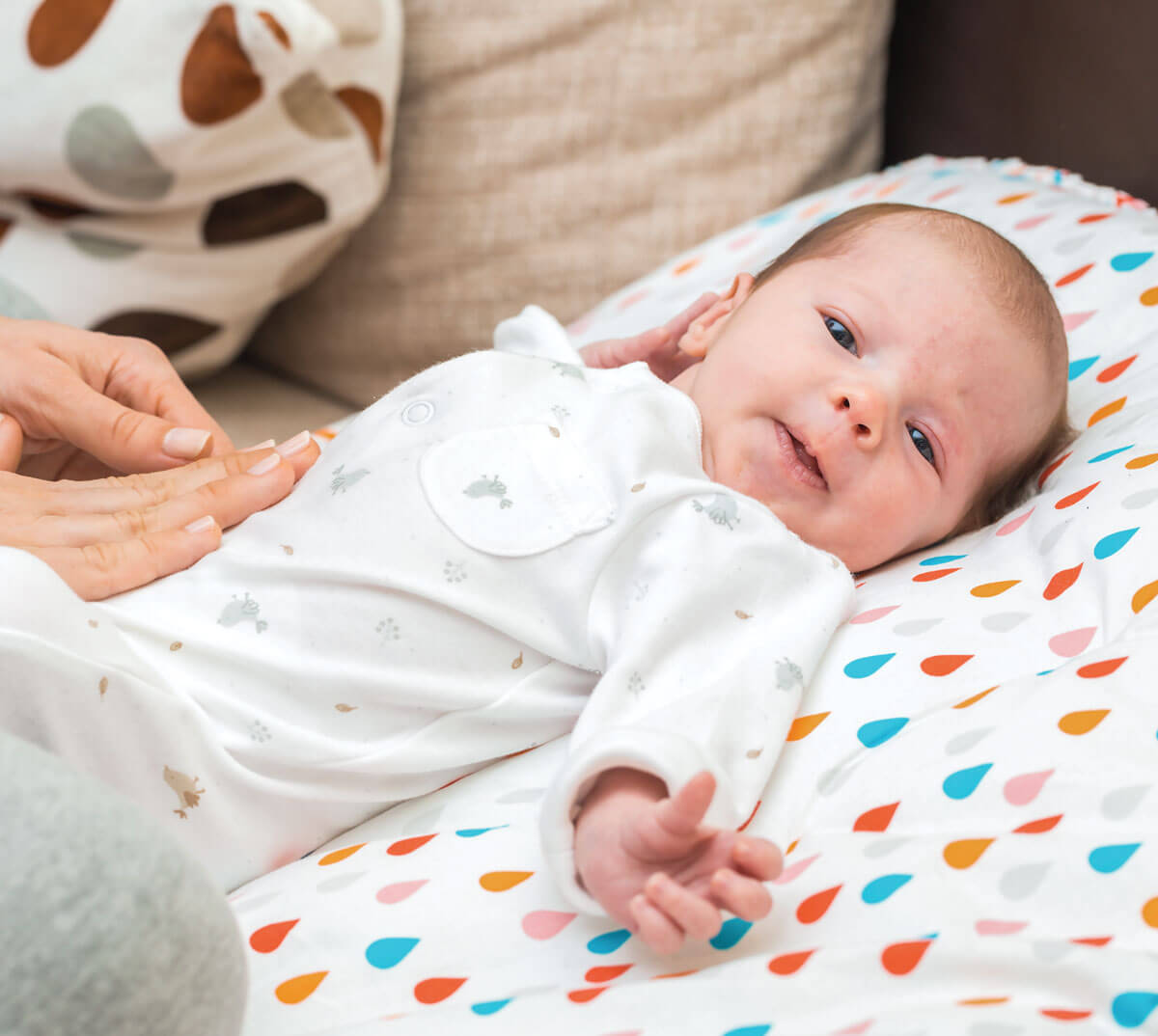 Baby smiling, lying on colorful blanket, wearing white onesie.