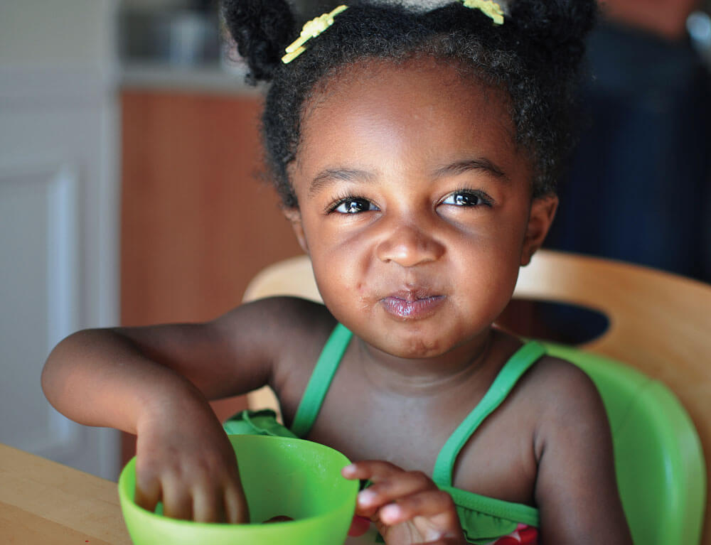 Child enjoying snack in a green bowl, smiling brightly.