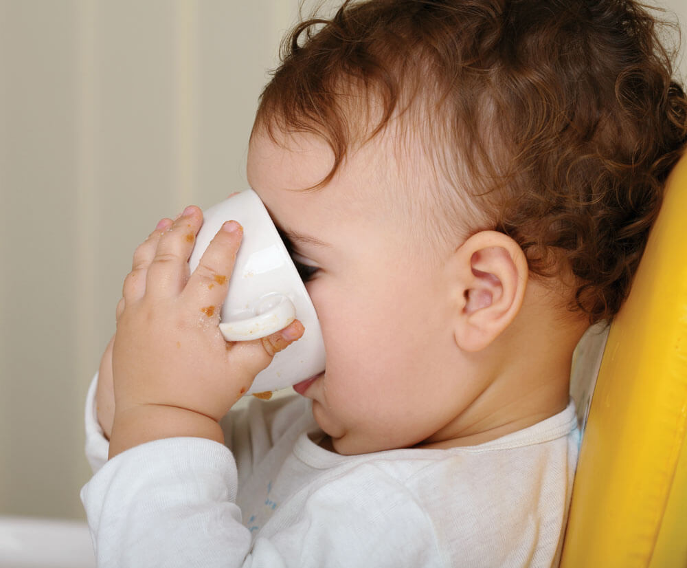 Toddler drinking from cup, sitting on chair.