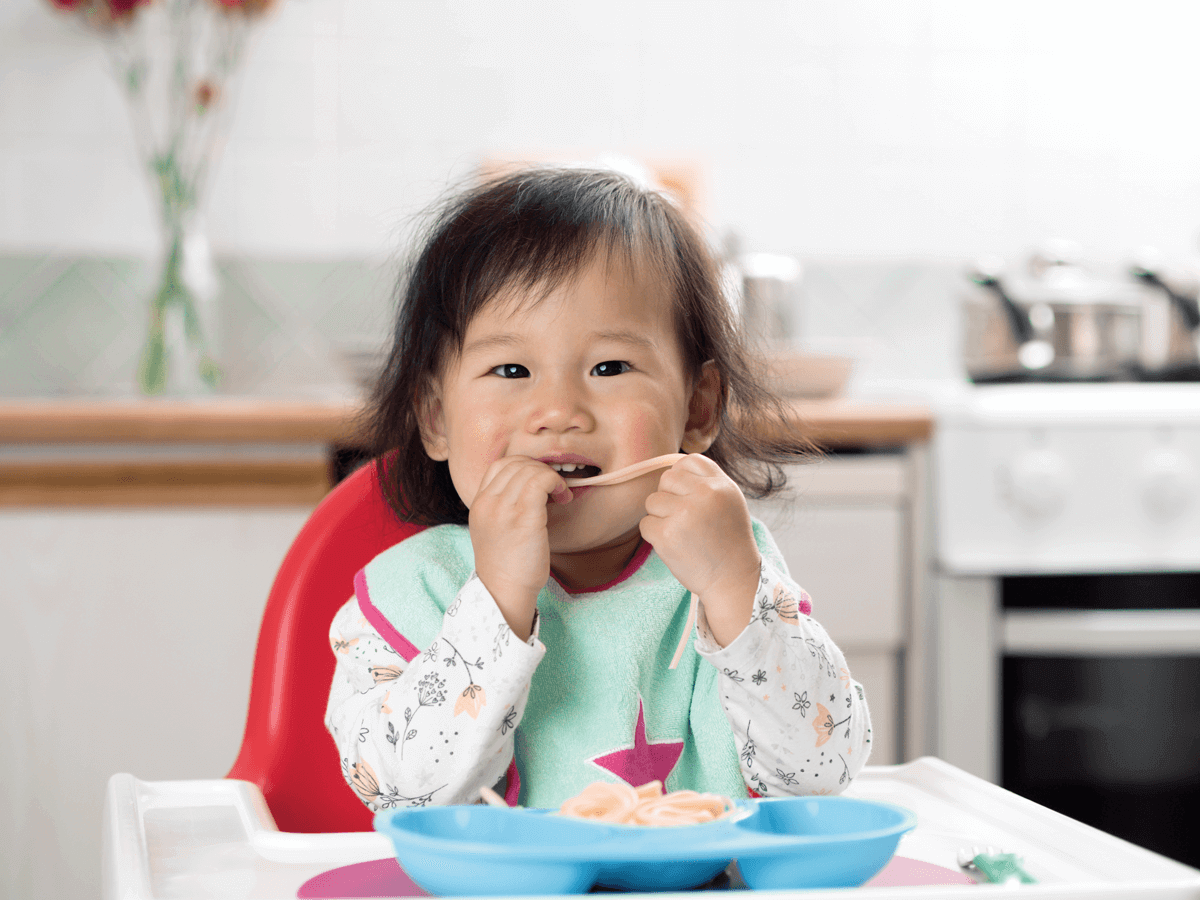 Toddler joyfully eating pasta at kitchen table.