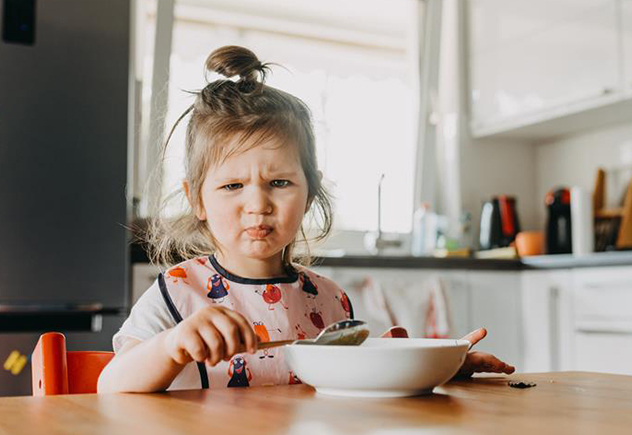 Child frowning while sitting with a bowl in kitchen.