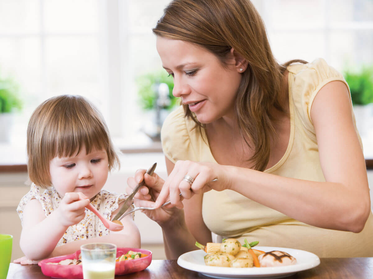 Mother helping young child eat with utensils.