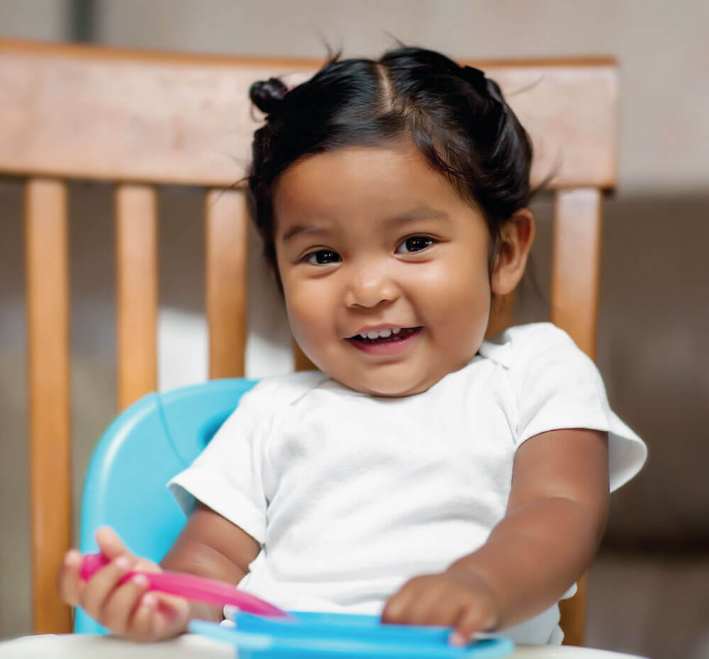 Smiling toddler holding utensils in high chair.