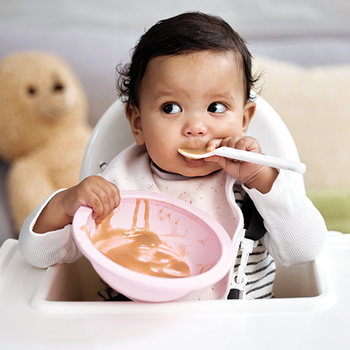 Baby eating with spoon from a pink bowl.