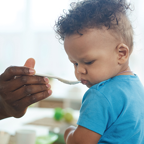 Infant in blue shirt being spoon-fed gently.