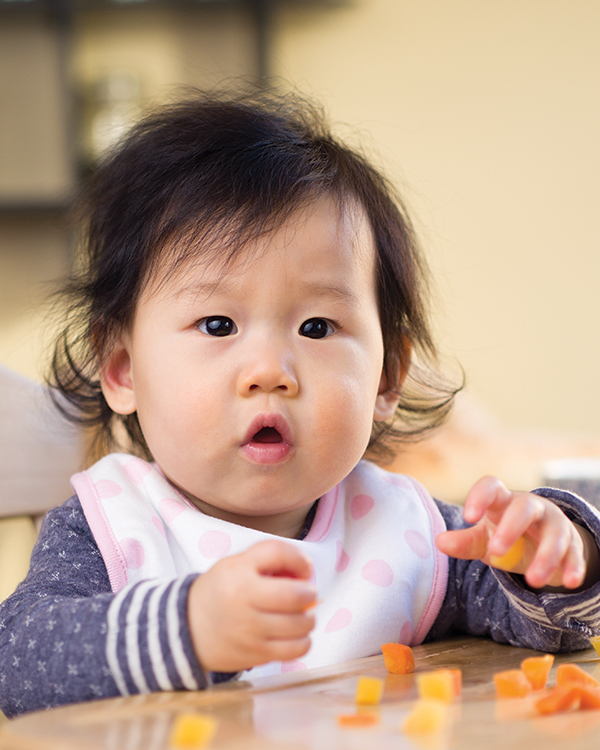 Baby eating small cubes of food on table.