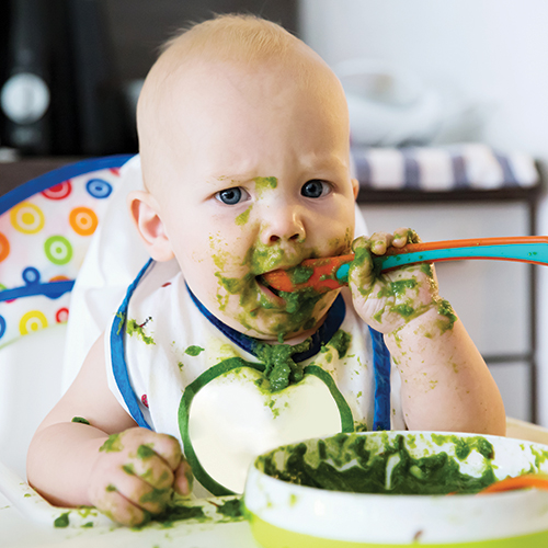 Baby eating messily with green food everywhere.