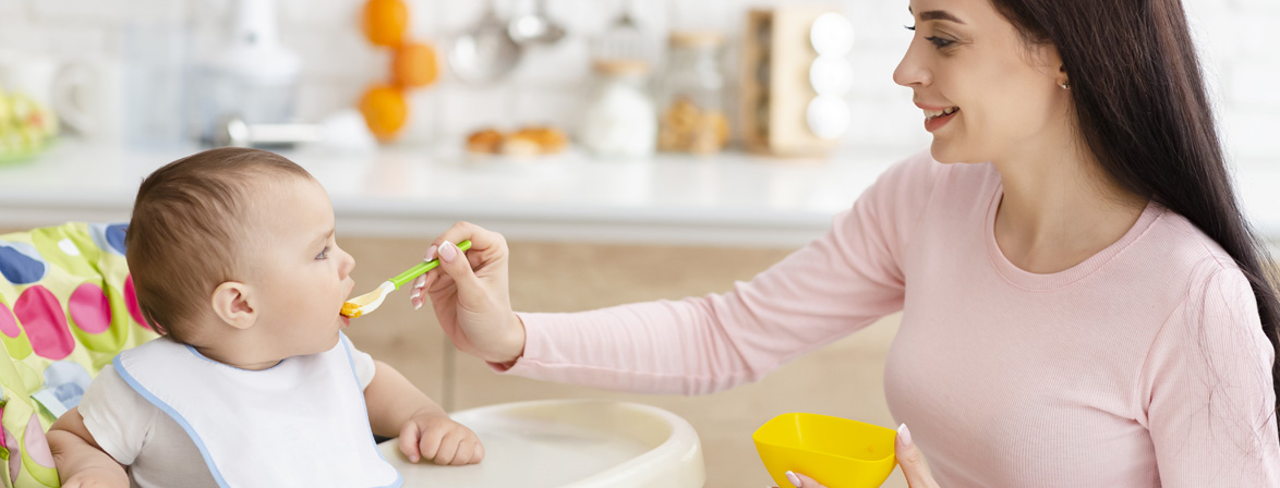 Mother feeds baby in bright kitchen setting.