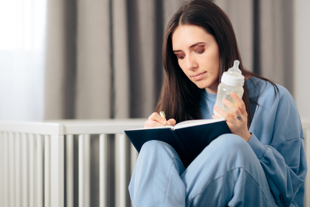 Woman writing in notebook, holding baby bottle.