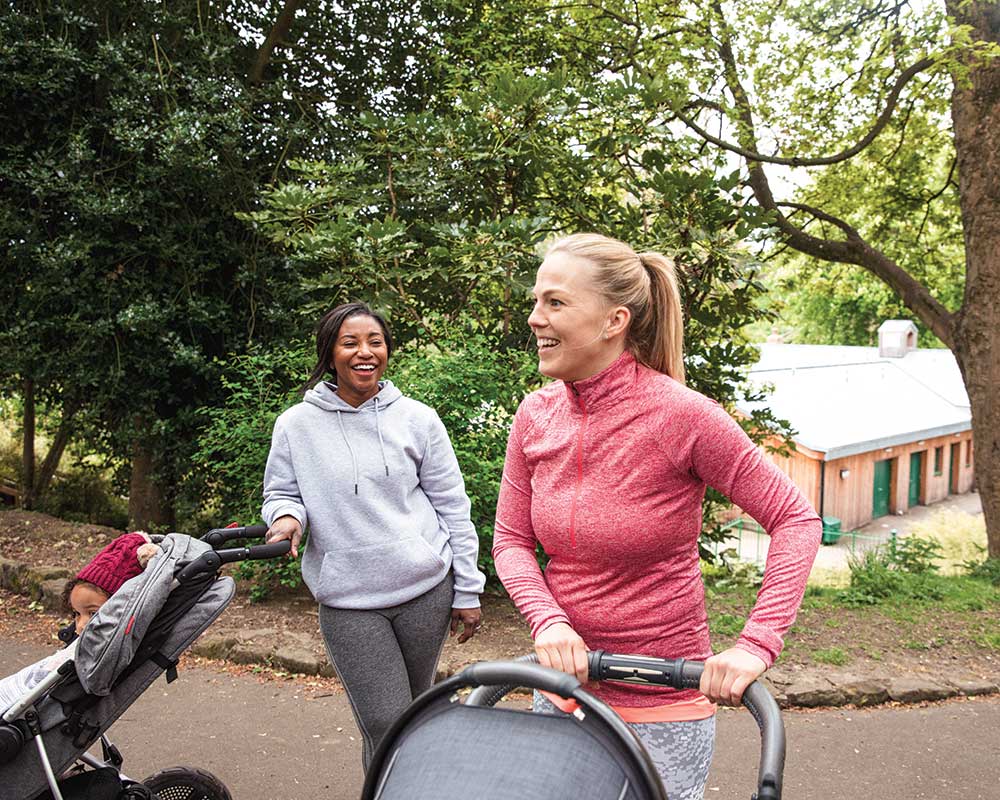 Two women smiling, walking outdoors with strollers.