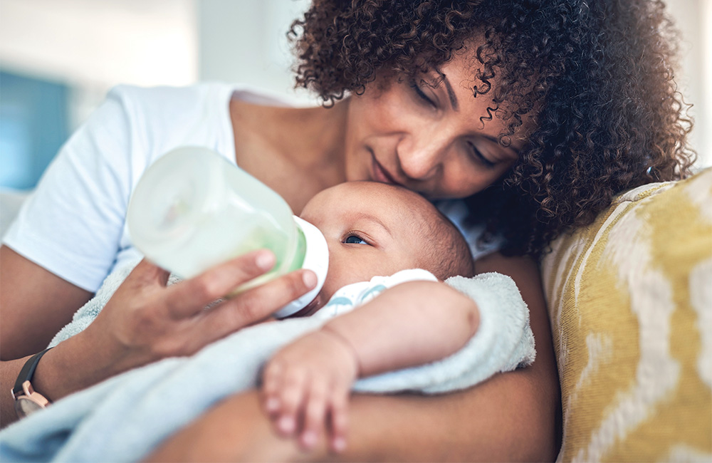 Mother lovingly feeds baby with bottle on couch.