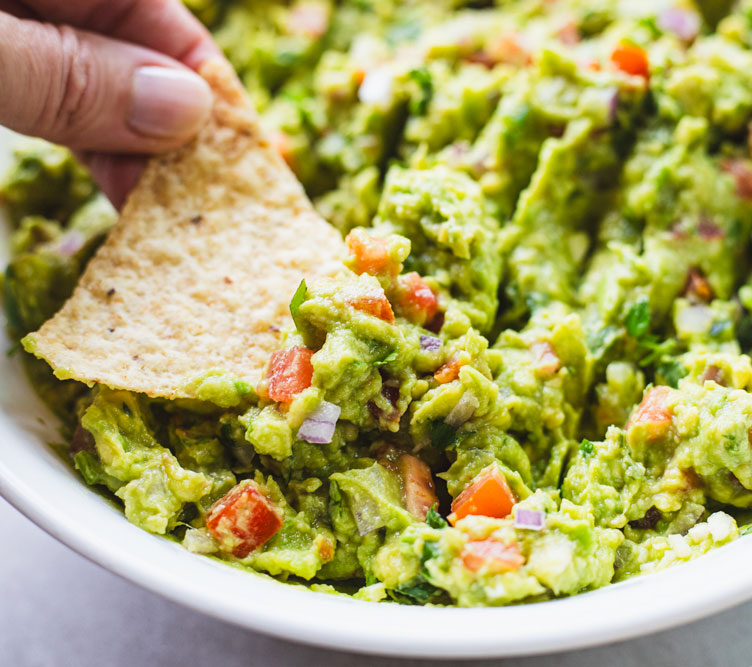 Hand dips chip into fresh guacamole with tomatoes.