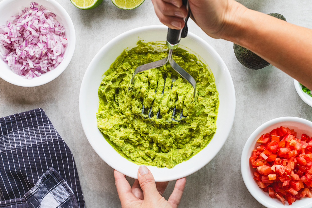 Hands mashing avocados in bowl, surrounded by ingredients.