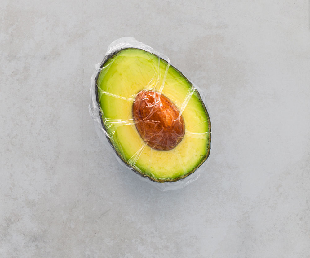 Half avocado wrapped in clear plastic on table.
