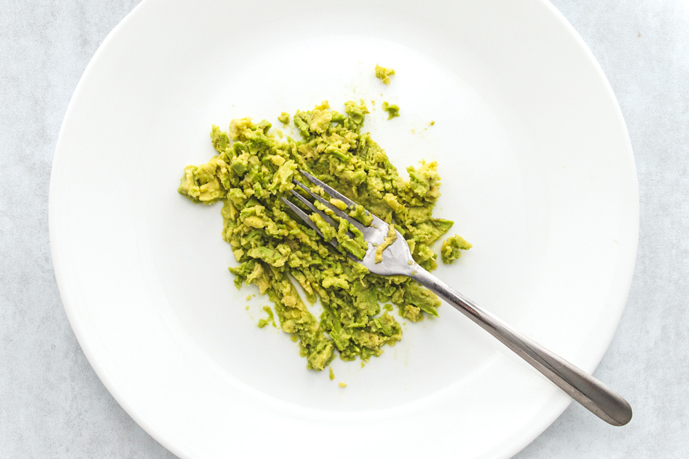 Mashed avocado with fork on white plate.