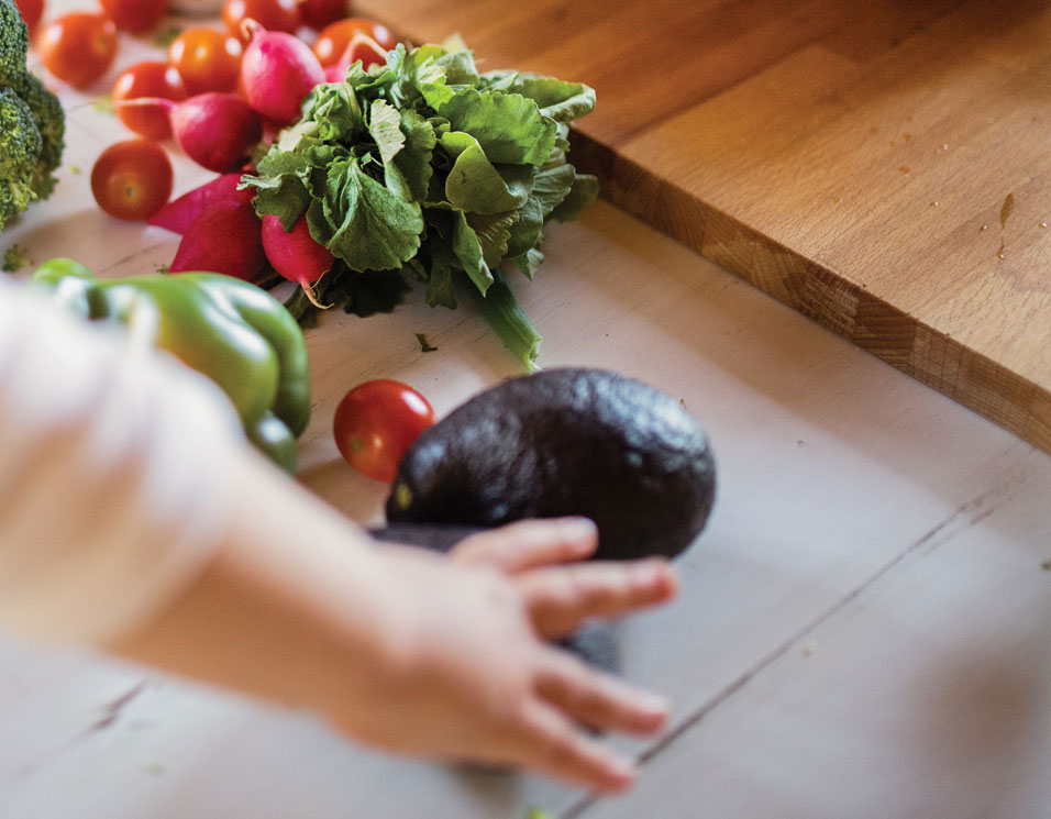 Child's hand reaching for fresh vegetables on table.