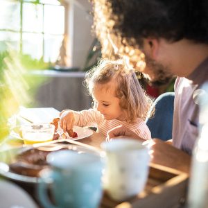 Child eating breakfast with adult at sunny table.