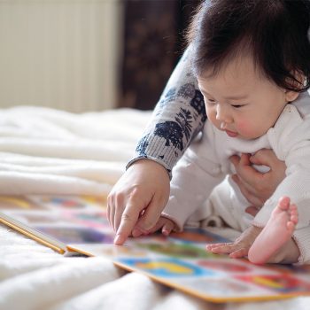 baby-learning Baby and adult reading colorful book together on bed.